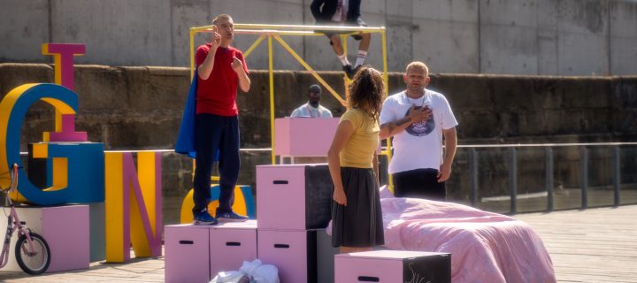 photo of 4 people performing in a play outdoors. a man looks at a young girl who is turning to face him. Another person stands behind watching. there are pink blocks structure surrounding them.