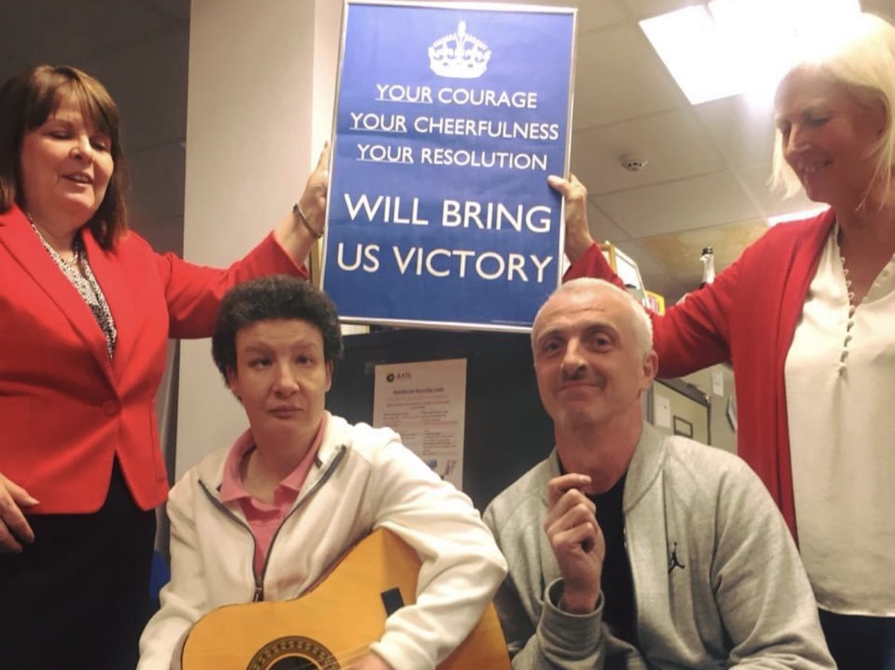 Photo of 2 women holding a sign reading 'Your courage, your cheerfulness, your resolution will bring us victory' in front is a woman playing a guitar and a man smiling. 