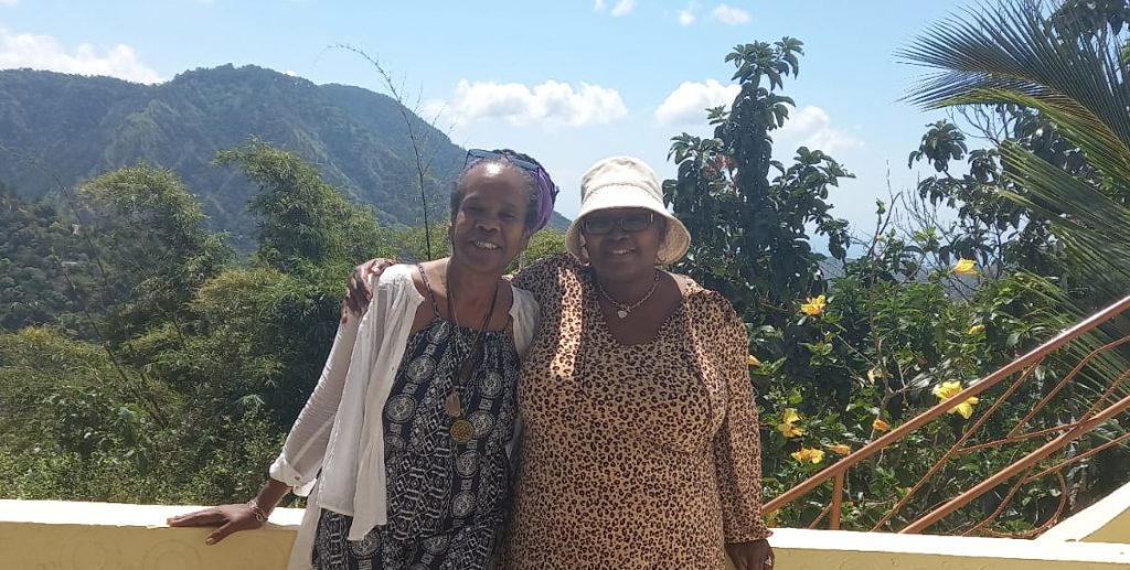 photo of two women posing and smiling with the backdrop of mountains in Jamaica. The women are middle aged and black wearing summer dresses. One wears a hat